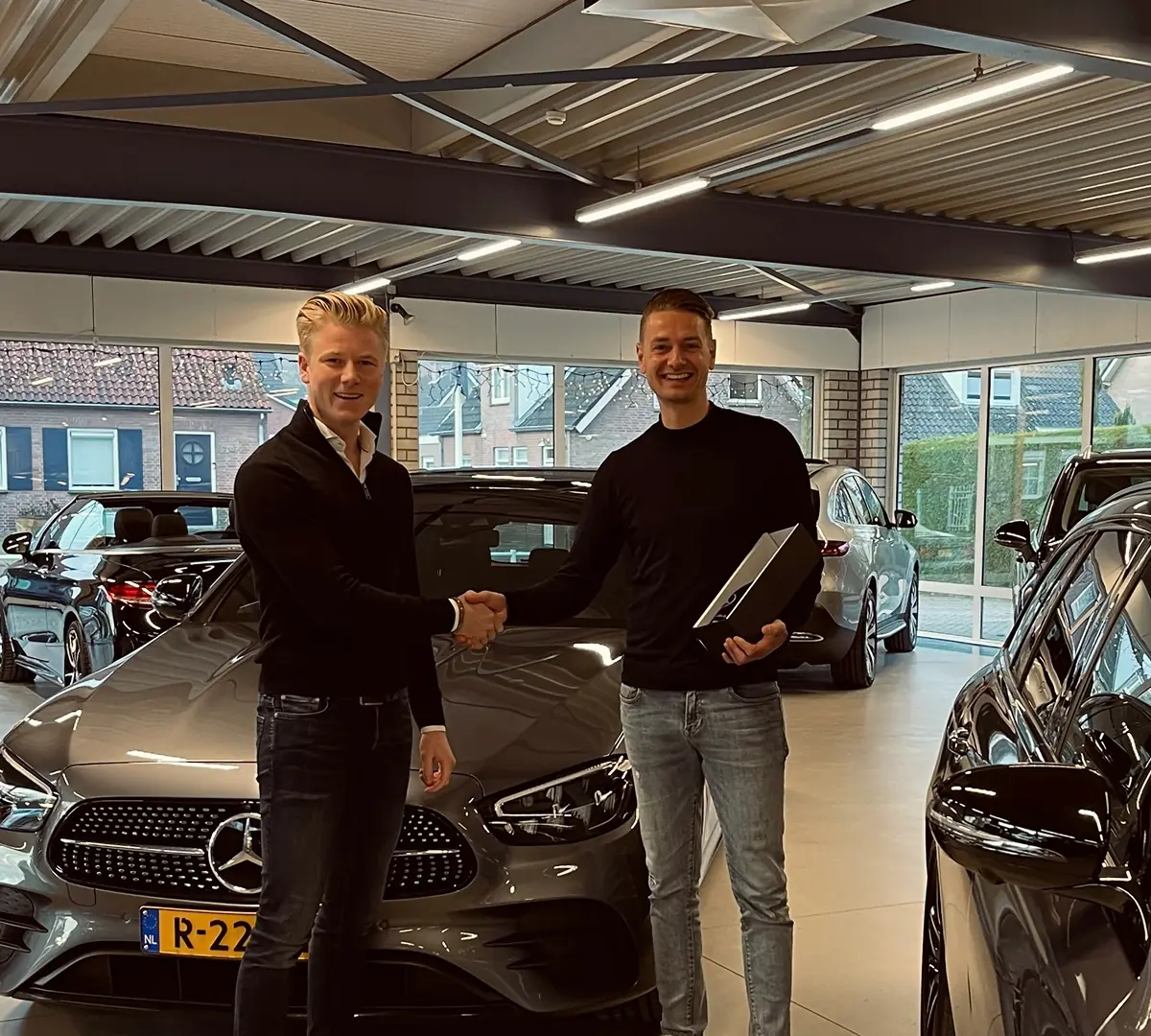 Twee mannen schudden handen bij een grijze Mercedes-Benz in een autoshowroom.